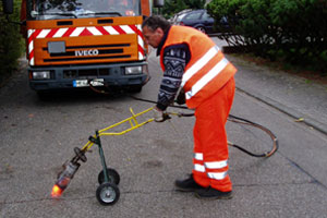 Ein Arbeiter in orangefarbener Schutzkleidung verwendet einen Schweißbrenner auf Rädern, um einen Straßenbelag zu erhitzen oder zu reparieren, während hinter ihm ein orangefarbener Lastwagen parkt.