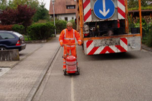 Ein Arbeiter in einem orangefarbenen Sicherheitsanzug malt mit einer Markierungsmaschine eine Linie auf eine Straße, während ein Lkw mit einem großen blauen Pfeil vorausfährt. Im Hintergrund sind Bäume und Häuser zu sehen.