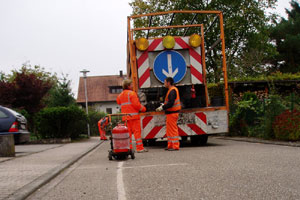Zwei Straßenarbeiter in orangefarbener Sicherheitskleidung stehen hinter einem Arbeitsfahrzeug mit einem blauen Pfeil, das in einer Wohnstraße steht. Neben ihnen steht ein Feuerlöscher auf der Straße. Im Hintergrund sind Bäume und Häuser zu sehen.
