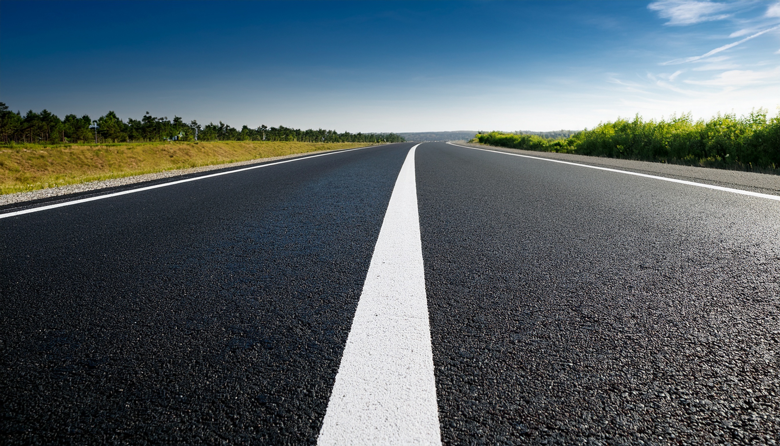 Eine frisch geteerte, leere Asphaltstraße mit weißen Fahrbahnmarkierungen erstreckt sich unter einem strahlend blauen Himmel in die Ferne, gesäumt von Grünflächen und Bäumen auf beiden Seiten.