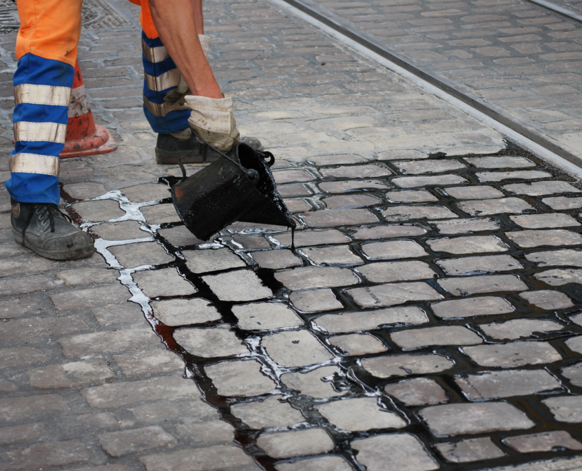 Ein Arbeiter in orange-blauen Sicherheitshosen schüttet schwarzen Teer aus einem Container auf das Kopfsteinpflaster, um Risse zwischen den Steinen in der Nähe der Straßenbahnschienen zu versiegeln.