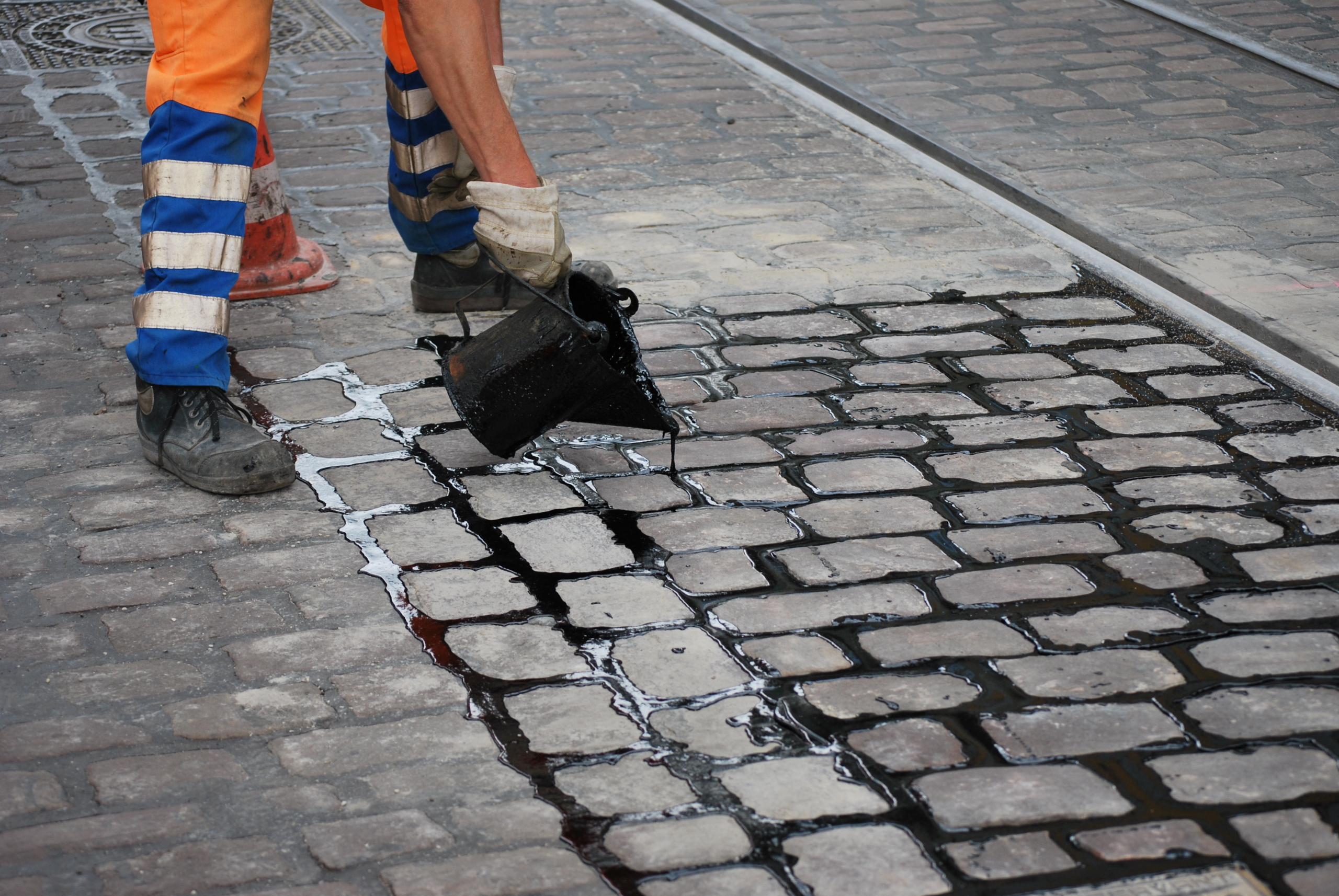 Ein Arbeiter in orange-blauen Sicherheitshosen schüttet schwarzen Teer aus einem Container auf das Kopfsteinpflaster, um Risse zwischen den Steinen in der Nähe der Straßenbahnschienen zu versiegeln.