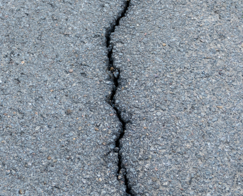 Eine Nahaufnahme einer rissigen Asphaltstraße mit einem gezackten, dunklen Riss, der sich senkrecht durch die Mitte der rauen, grauen Oberfläche zieht.