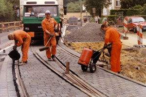 Drei Bauarbeiter in orangefarbenen Overalls reparieren oder verlegen Straßenbahnschienen auf einer Kopfsteinpflasterstraße. Einer fegt, ein anderer setzt eine Maschine ein, und ein dritter schiebt Geräte, während im Hintergrund ein Lkw und Absperrungen zu sehen sind.