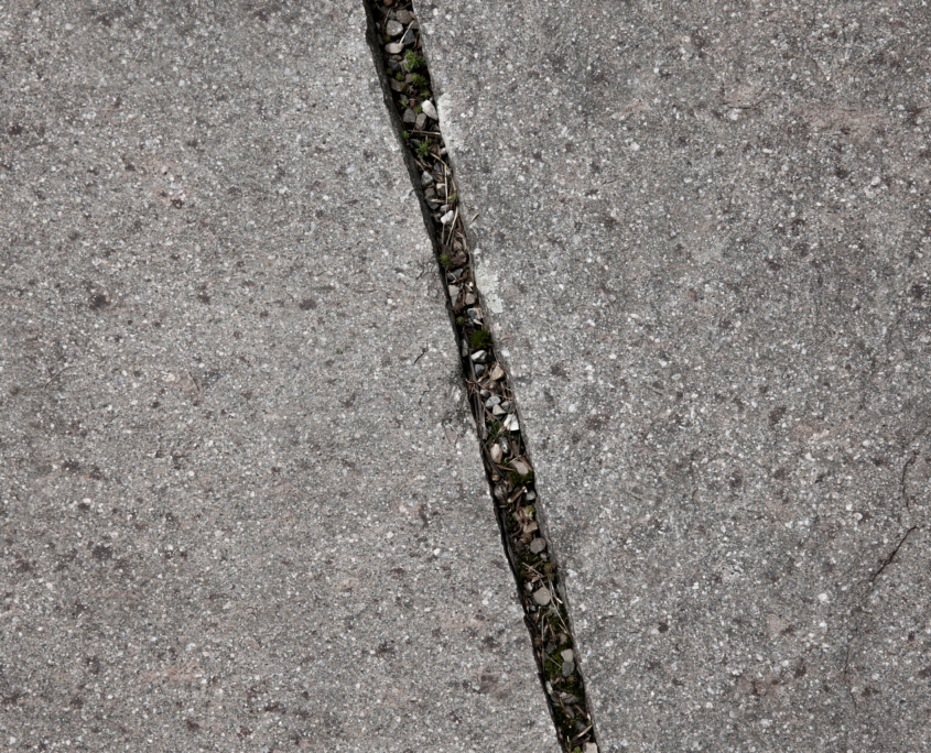 Nahaufnahme einer rissigen Betonoberfläche mit kleinen grünen Pflanzen und Schutt, der in dem schmalen Spalt wächst.