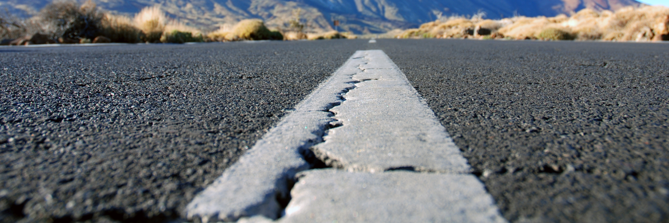 Nahaufnahme einer rissigen weißen Linie auf einer Asphaltstraße, mit trockenem Gras und Bergen im Hintergrund, die an eine Wüste oder trockene Landschaft erinnern. Das Foto ist aus einer niedrigen, ebenerdigen Perspektive aufgenommen.