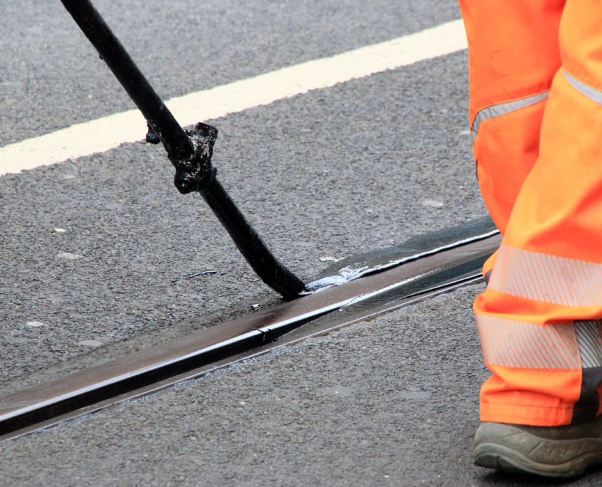 Ein Arbeiter in leuchtend orangen Hosen trägt mit einem langstieligen Werkzeug schwarze Dichtungsmasse auf einen Riss in der Asphaltstraße auf. Im Hintergrund ist eine gelbe Straßenmarkierung zu sehen.