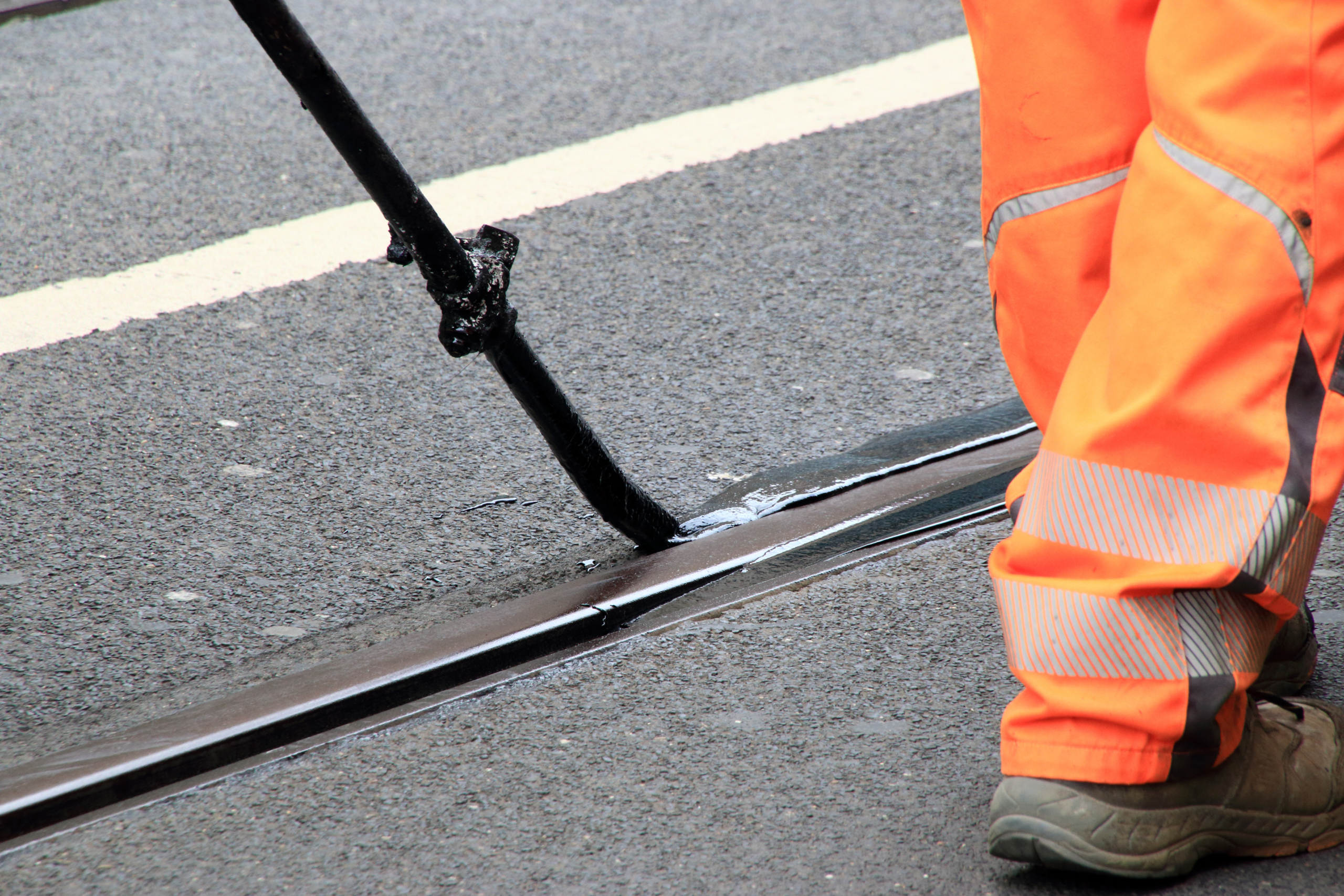 Ein Arbeiter in leuchtend orangen Hosen trägt mit einem langstieligen Werkzeug schwarze Dichtungsmasse auf einen Riss in der Asphaltstraße auf. Im Hintergrund ist eine gelbe Straßenmarkierung zu sehen.